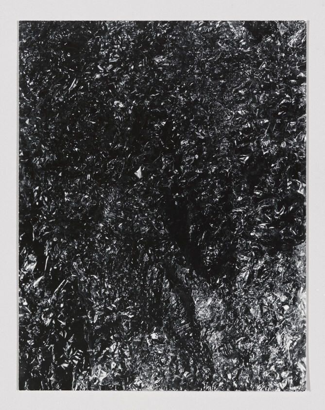 A monochrome photograph displaying a textured surface that resembles crumpled foil or a metallic, reflective material with intricate light and shadow interplay.