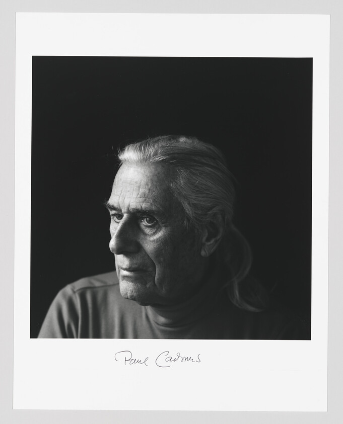 Older person with long hair pulled back looks thoughtfully to the left against a dark background.