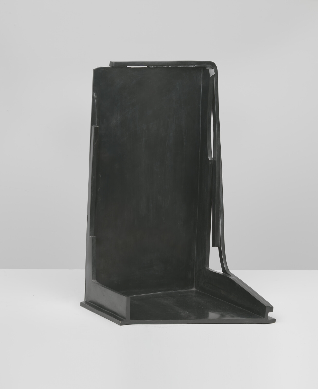 Tall dark abstract metal sculpture with angular vertical back and slanted base standing on white surface.