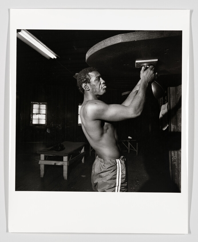 Shirtless man practices punching a speed bag in a dimly lit gym.