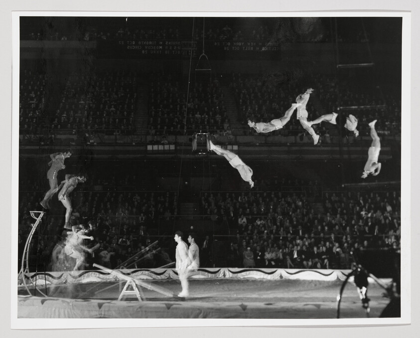 Acrobats perform multiple airborne somersaults over a trampoline in front of a seated audience.