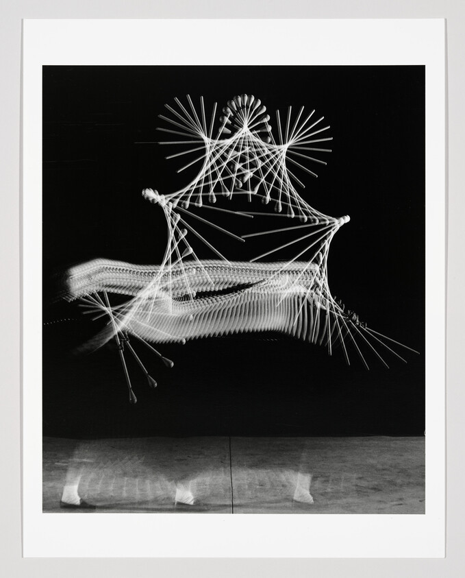 Black and white photograph capturing a long exposure of a Ferris wheel in motion, resulting in light trails that create a dynamic, abstract pattern against a dark background. The image is framed with a white border.