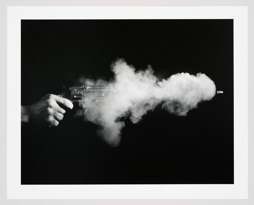 A hand fires a revolver, producing a cloud of smoke and a visible bullet exiting the barrel.