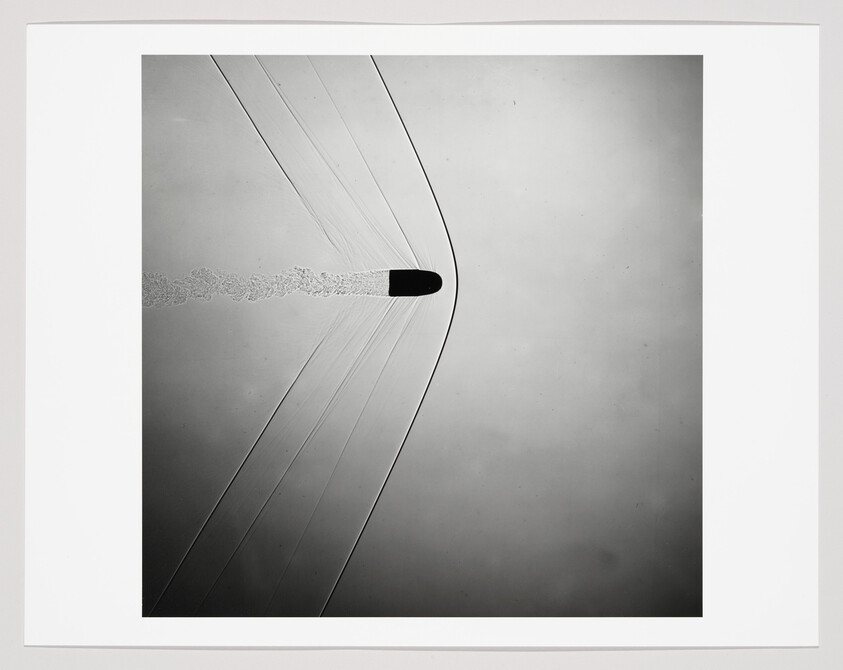 Bullet piercing a thin membrane, leaving curved shock lines and a trailing cloud.