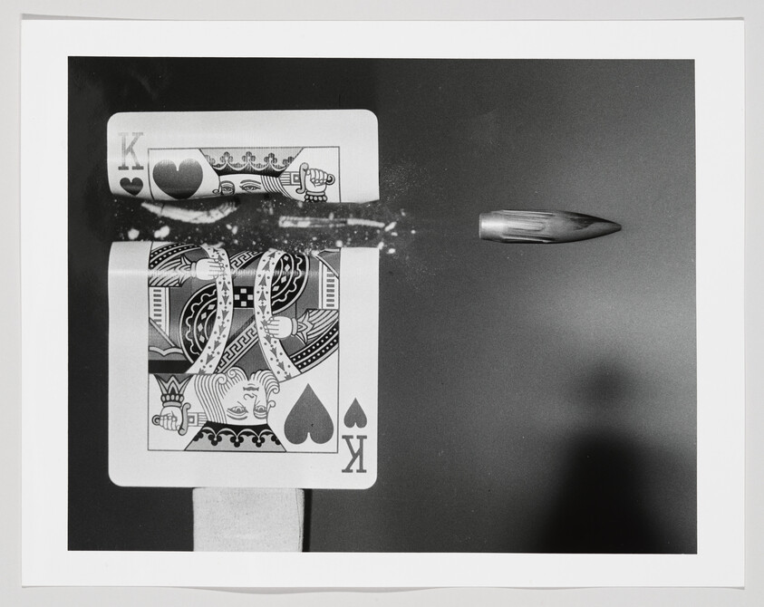 A bullet slices through a king playing card, sending paper fragments and dust into the air.