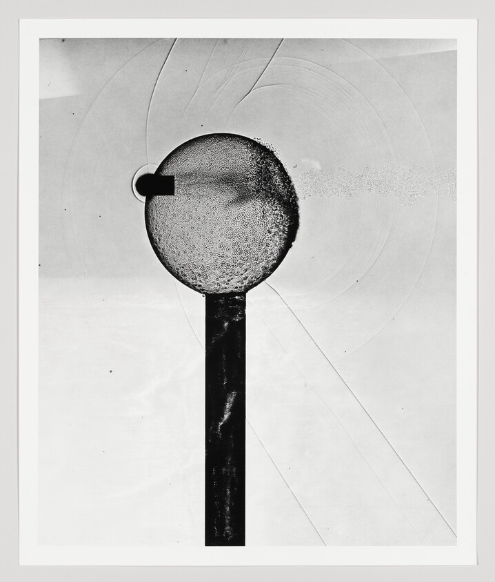 A bullet pierces a soap bubble on a vertical stand, spraying droplets to the right.