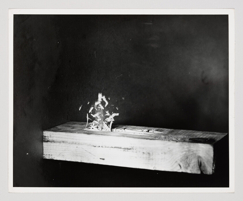 A wooden plank with wood shavings burning and sparks rising against a dark background.