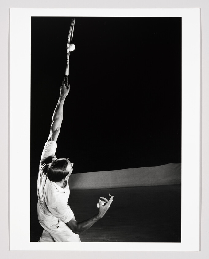 A tennis player reaches up with racket to serve as the ball hovers above.