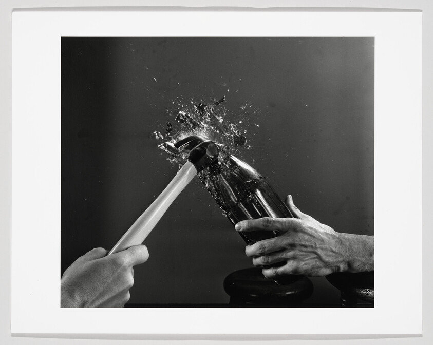 A hammer smashes a glass bottle held by a hand, sending shards and liquid flying.