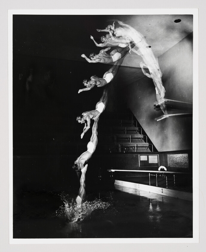 A black and white photograph capturing the motion of a diver in multiple stages of a dive, from the jump off the board to the entry into the water, creating a ghostly trail effect against the dark background of an indoor pool setting.