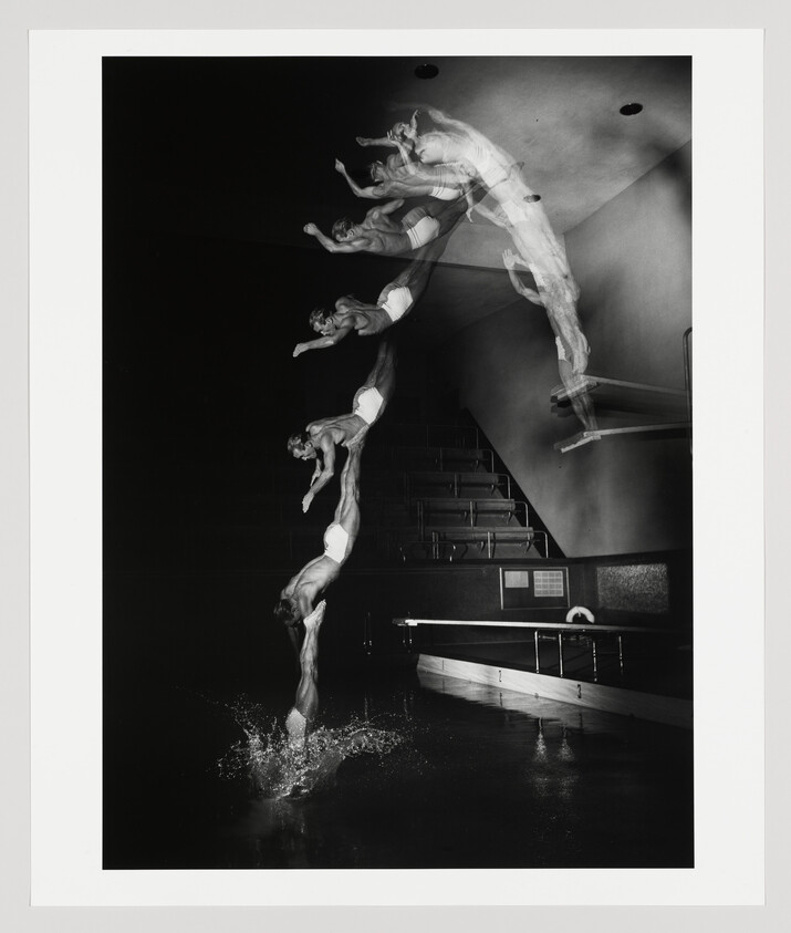 A black and white photograph capturing the motion of a diver in multiple stages of a dive, from the edge of a diving board to the splash upon entering the water, creating a ghostly trail of images against a dark indoor pool background.