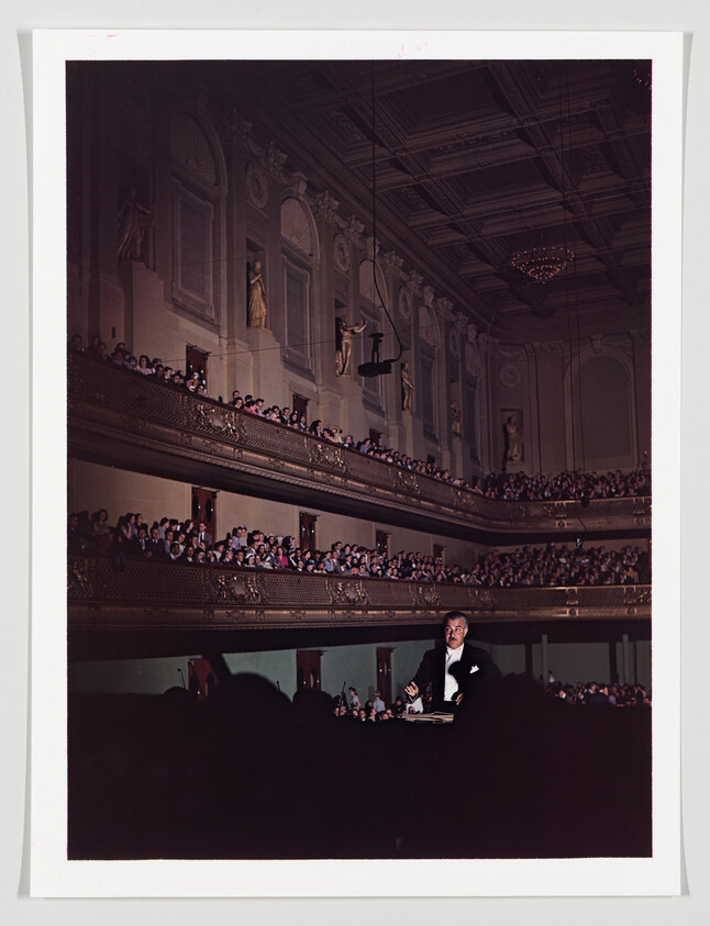 A conductor in a tuxedo leads a performance before crowded, ornate balconies in a concert hall.