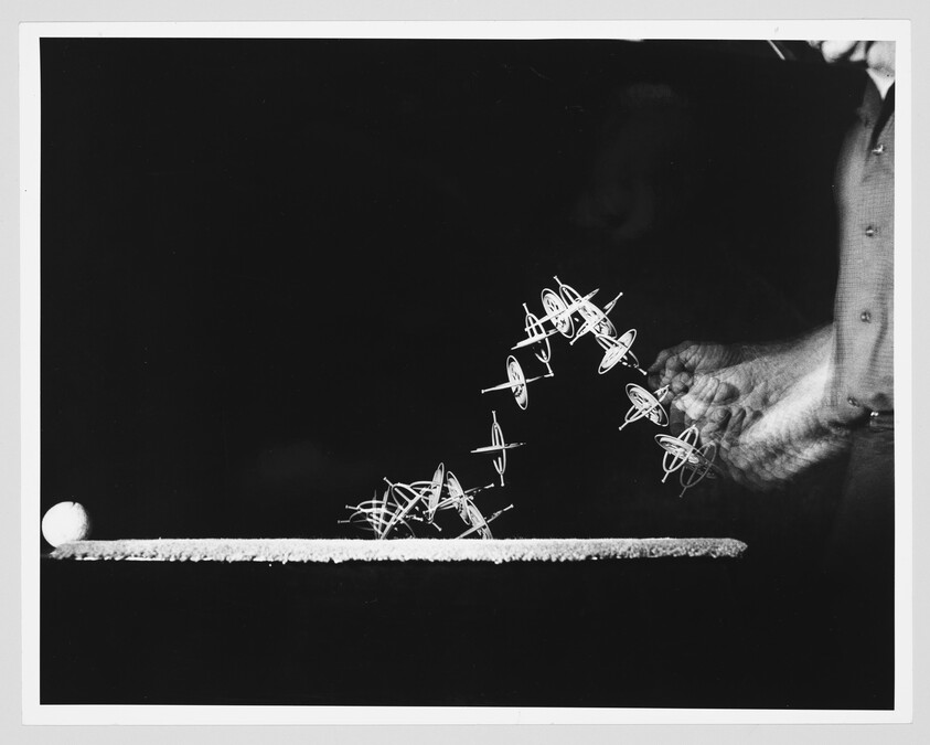 A hand flicks multiple spinning tops across a table, showing their curved motion path.