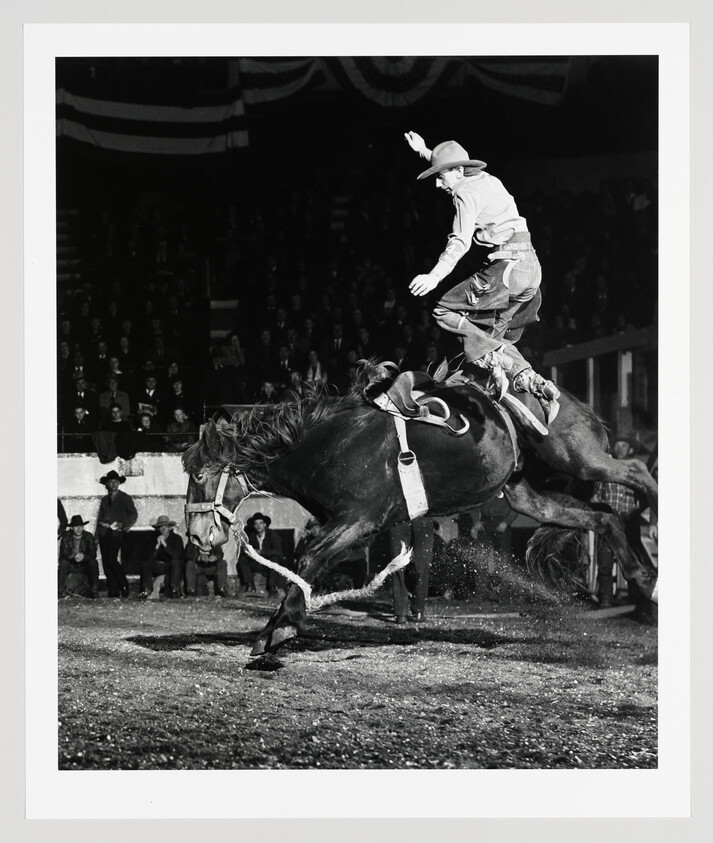 Cowboy in a hat rides a bucking horse at a packed rodeo, balancing with one arm raised.