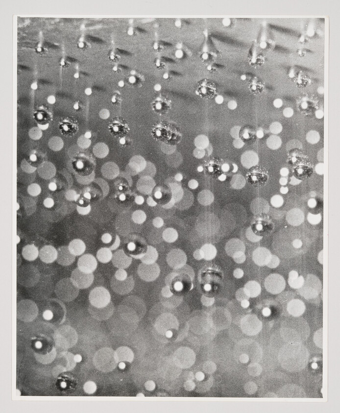 Close-up of hanging water droplets reflecting blurred circular lights in a black-and-white bokeh pattern.