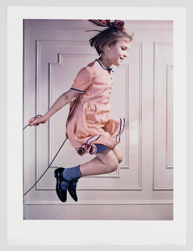 A young girl in a pink dress jumps rope midair against a paneled wall.