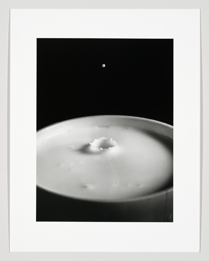 A droplet falls toward a cup of milk, creating a small crown splash on the surface.