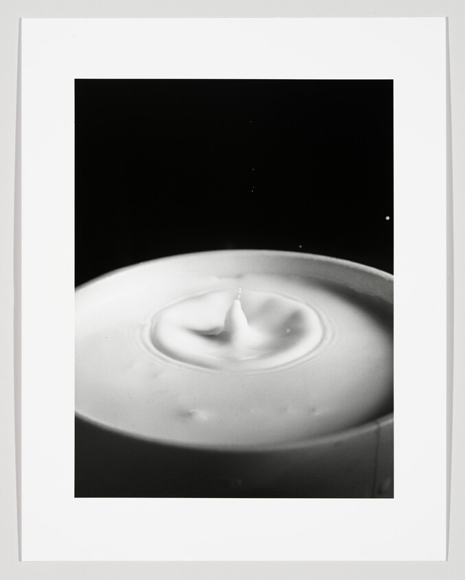 A high-contrast black and white photograph capturing a momentary milk droplet impact, creating a small, sharp peak and ripples on the surface of the milk. The image is framed with a white border.