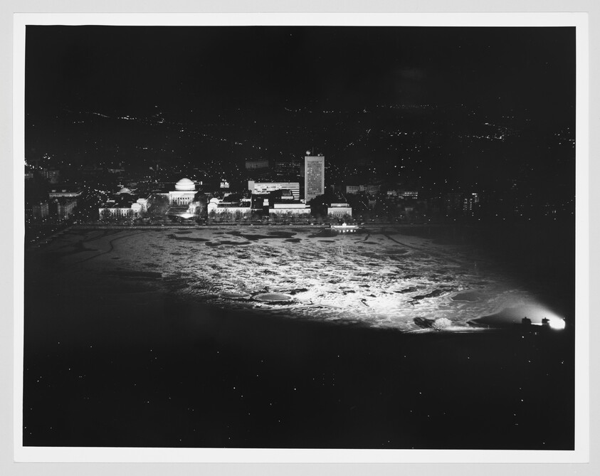 Aerial night view showing a frozen, spotlight-lit waterfront and illuminated city buildings across the bay.