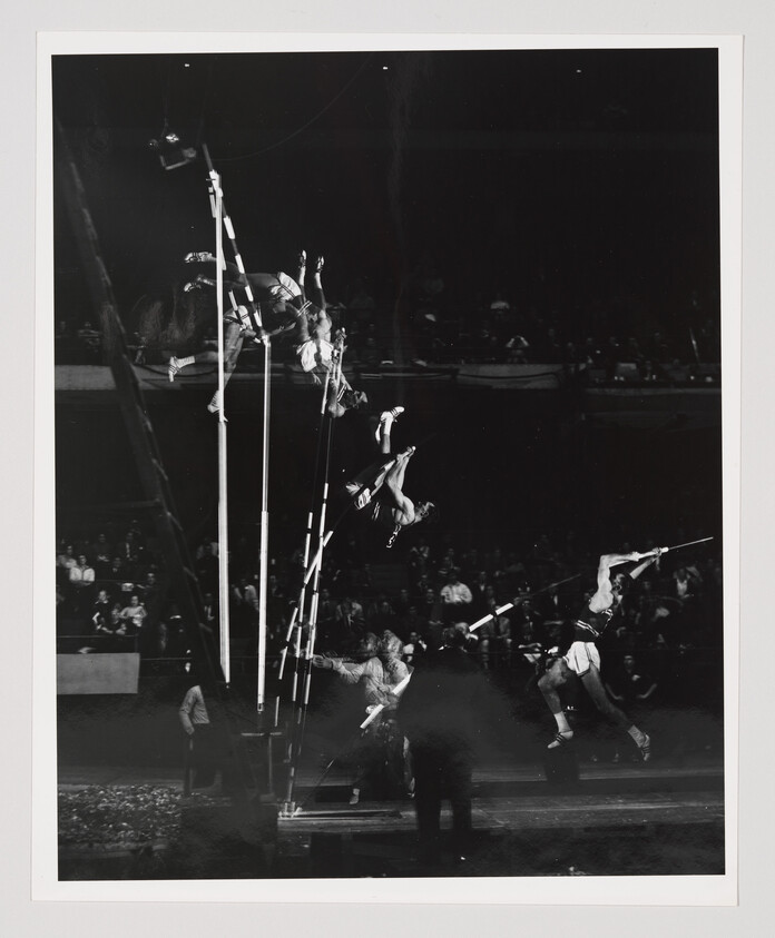 Several athletes perform pole vaulting and flips over poles in front of a seated audience.