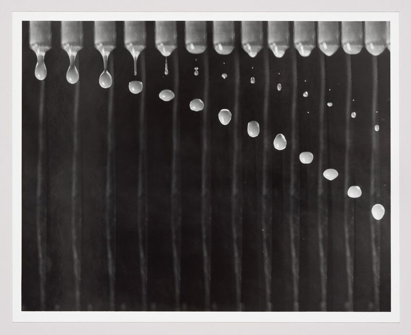 Row of metal nozzles releasing a diagonal trail of falling water droplets against a dark background.