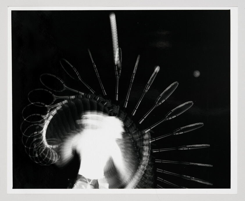 Black and white photograph capturing a motion blur of a spinning amusement park ride at night, with light trails creating circular patterns against a dark background.
