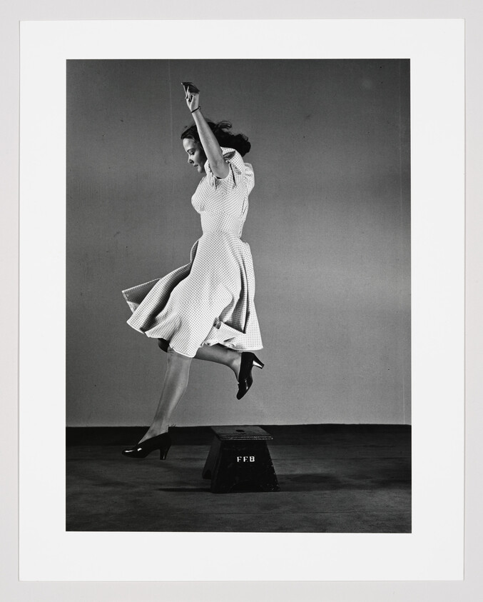 Woman in a polka-dot dress jumps off a small box marked "FFB," skirt billowing.