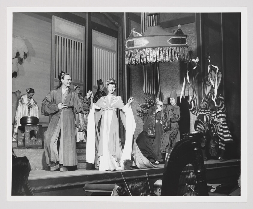 A stage production shows actors in traditional Asian costumes performing under a large hanging lantern.
