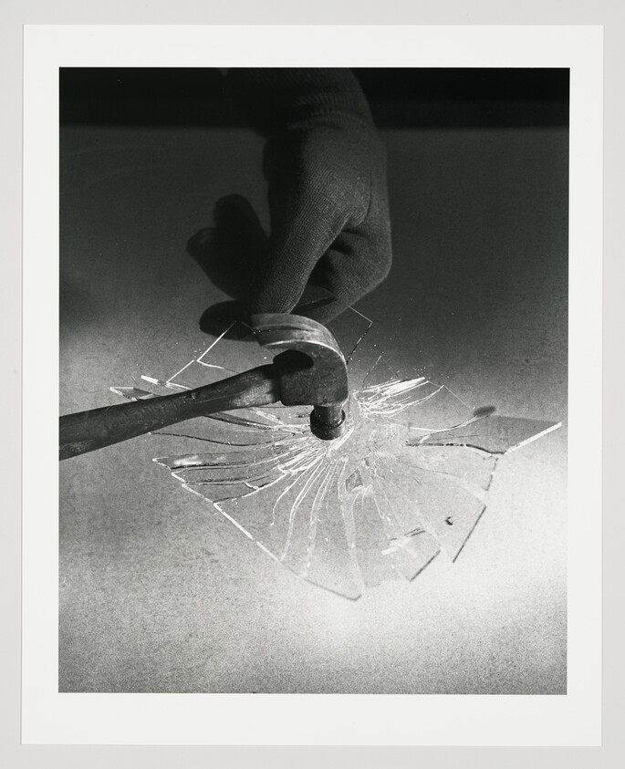 A gloved hand holds a hammer striking a glass pane, sending radial cracks outward.