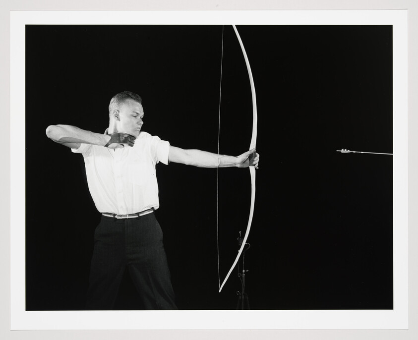 A man draws a longbow, holding it steady as an arrow flies to the right.