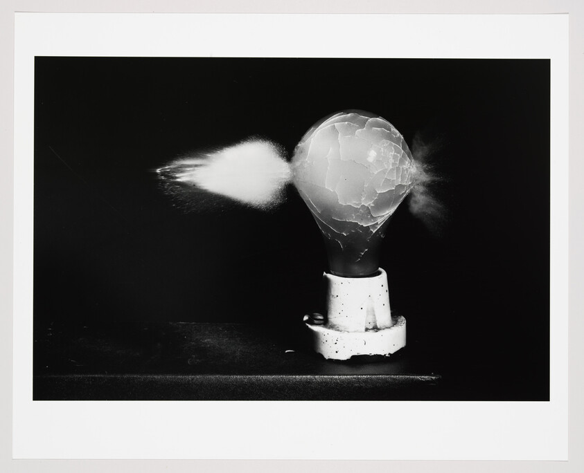 Light bulb shatters as a projectile passes through, sending glass fragments and dust outward.