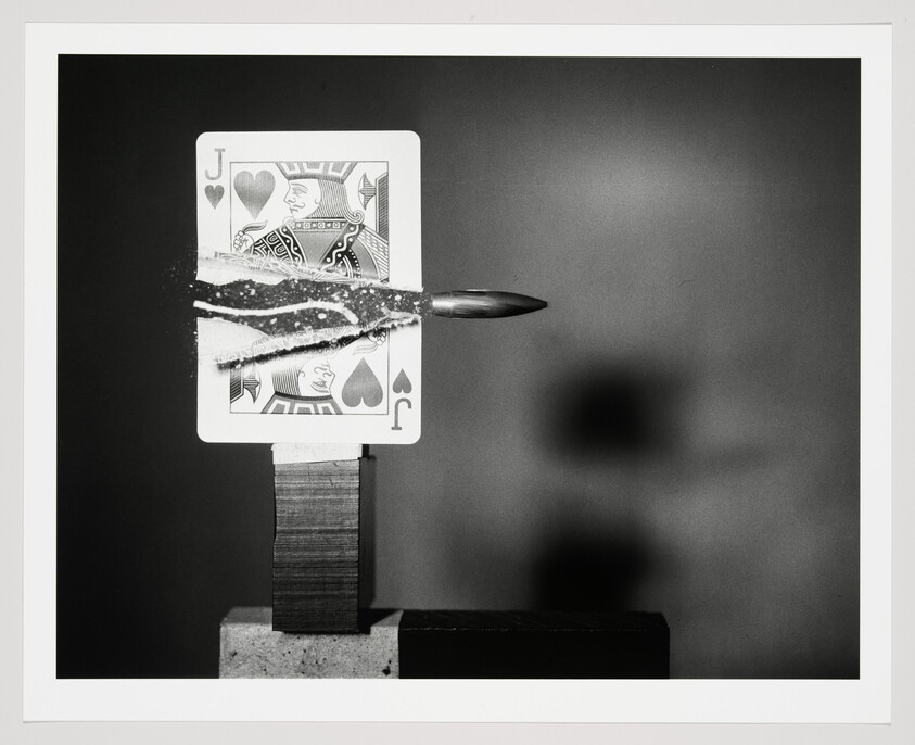 A bullet pierces a Jack of Hearts playing card, tearing paper and scattering fragments.