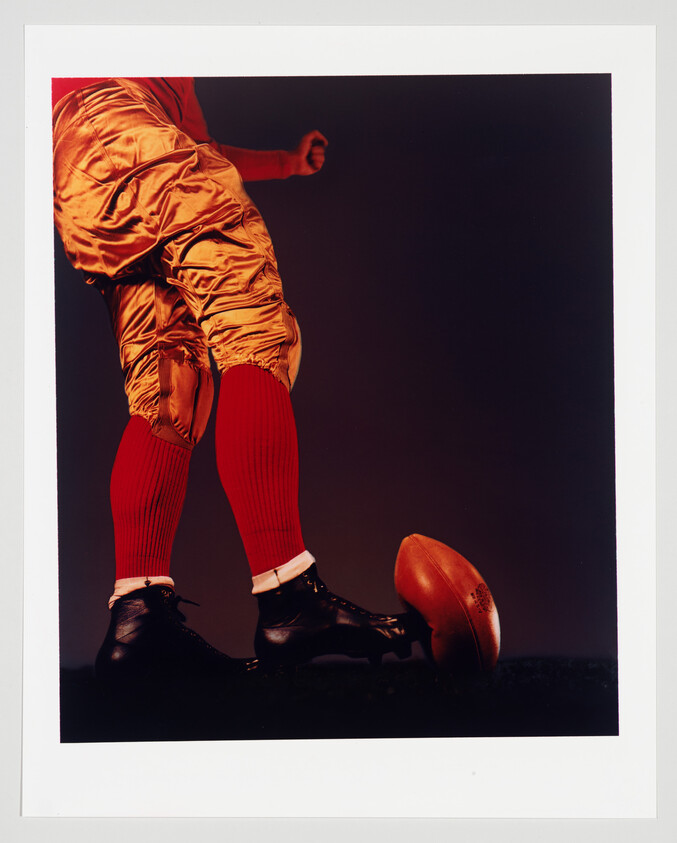 A close-up of a person's lower body dressed in American football gear with gold pants and red socks, kicking a football on a dark background.