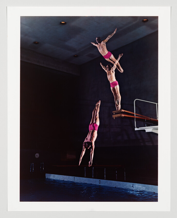 Three synchronized swimmers are captured in mid-air during a dive, with their positions staggered to create a cascading effect against the backdrop of an indoor swimming pool. The first swimmer is closest to the water, followed by the second and third who are progressively higher up. The image is well-lit, highlighting the swimmers' athletic forms and the calm surface of the pool below.