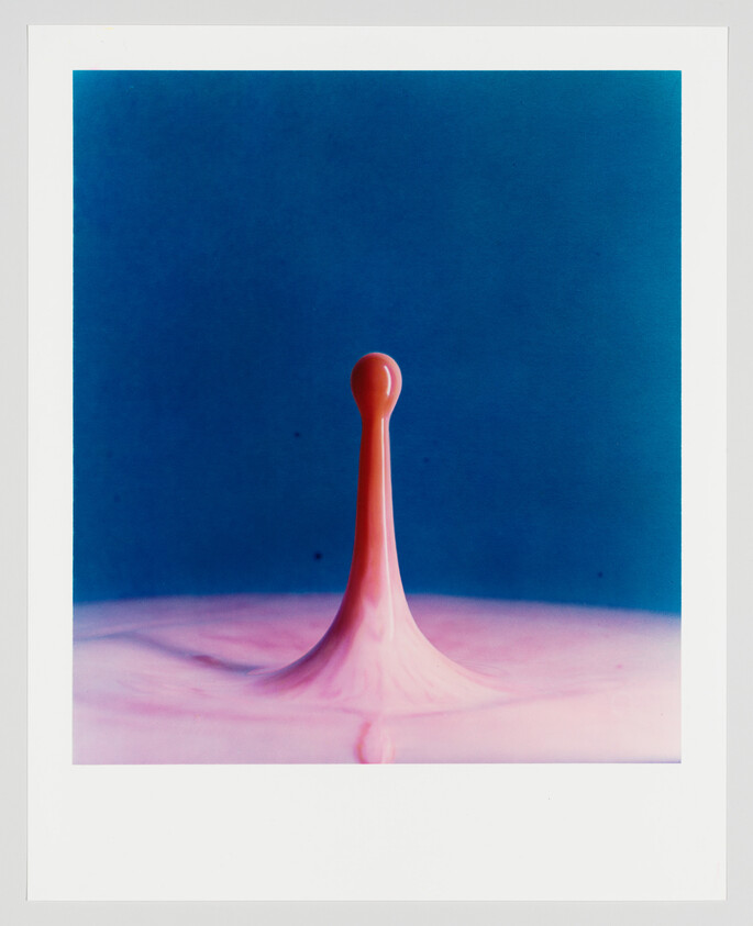 A high-speed photograph capturing a single drop of red liquid mid-splash against a solid blue background, creating a thin, elongated droplet shape above a rippled liquid surface.