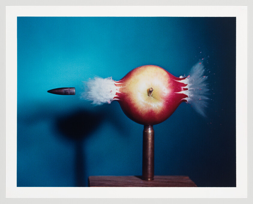 A high-speed photograph capturing the moment a bullet passes through an apple, which is mounted on a pedestal against a blue background. The bullet is entering from the left, with a visible white puff of impact, and apple fragments are explosively ejecting to the right.
