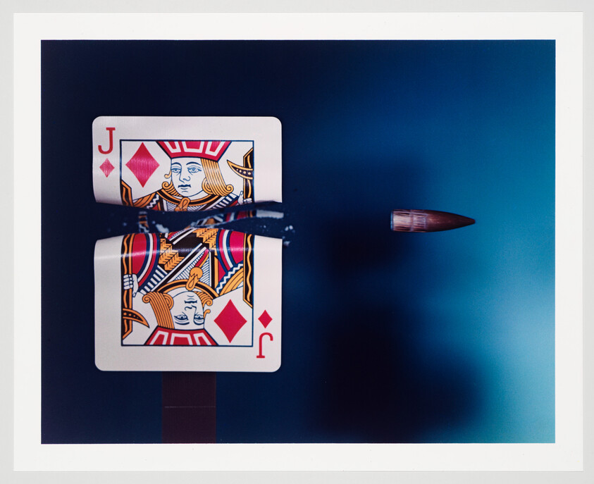 A Jack of Diamonds playing card is pierced by a bullet in mid-air against a dark blue background, with the bullet captured on the right side of the image showing motion blur. The card and bullet are reflected on a surface below, creating a symmetrical composition.