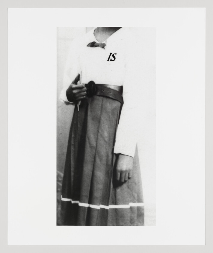 Young person wearing belted skirt and blouse with "IS" letters, holding a small book at their side.
