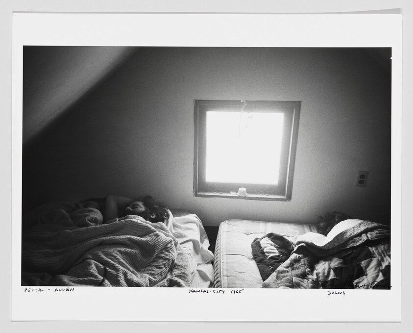 Two people sleep under blankets on separate mattresses in a dim attic room beneath a bright window.