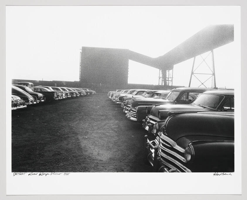 Rows of vintage cars parked in an industrial lot next to large conveyor structures.