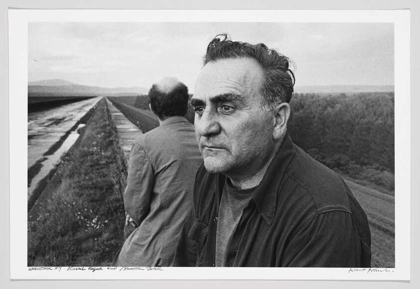 Two men stand beside a long canal, one facing the camera with a thoughtful, weathered expression.