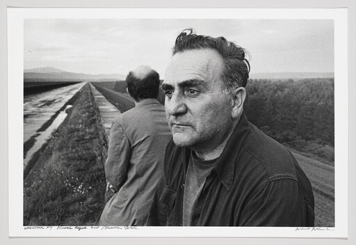 Two men stand beside a long canal, one facing the camera with a thoughtful, weathered expression.