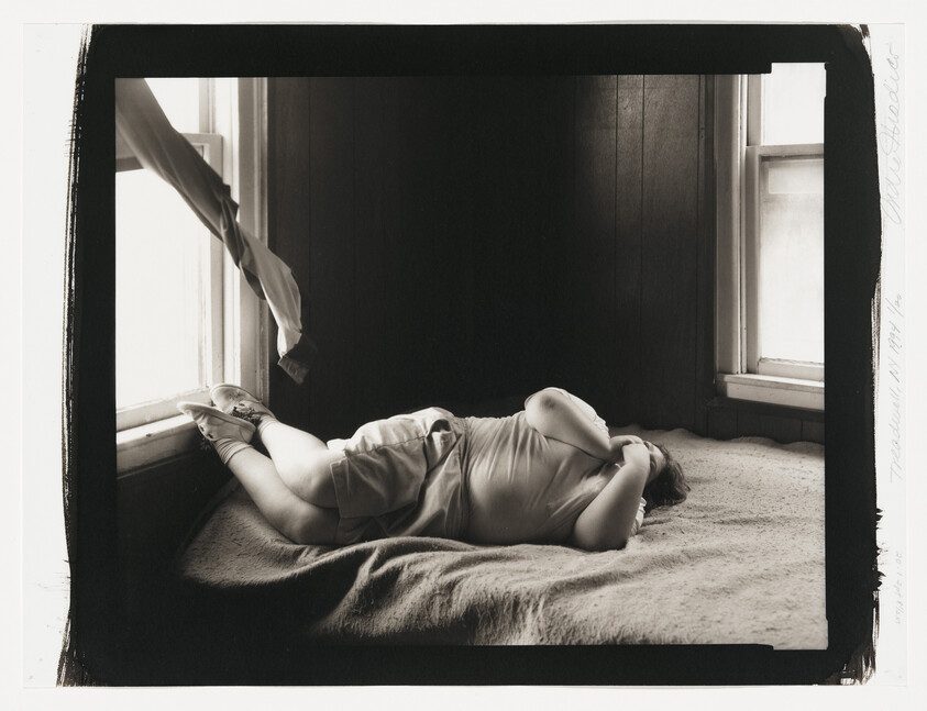 A person lies curled on a bed by two windows, hands covering their face.