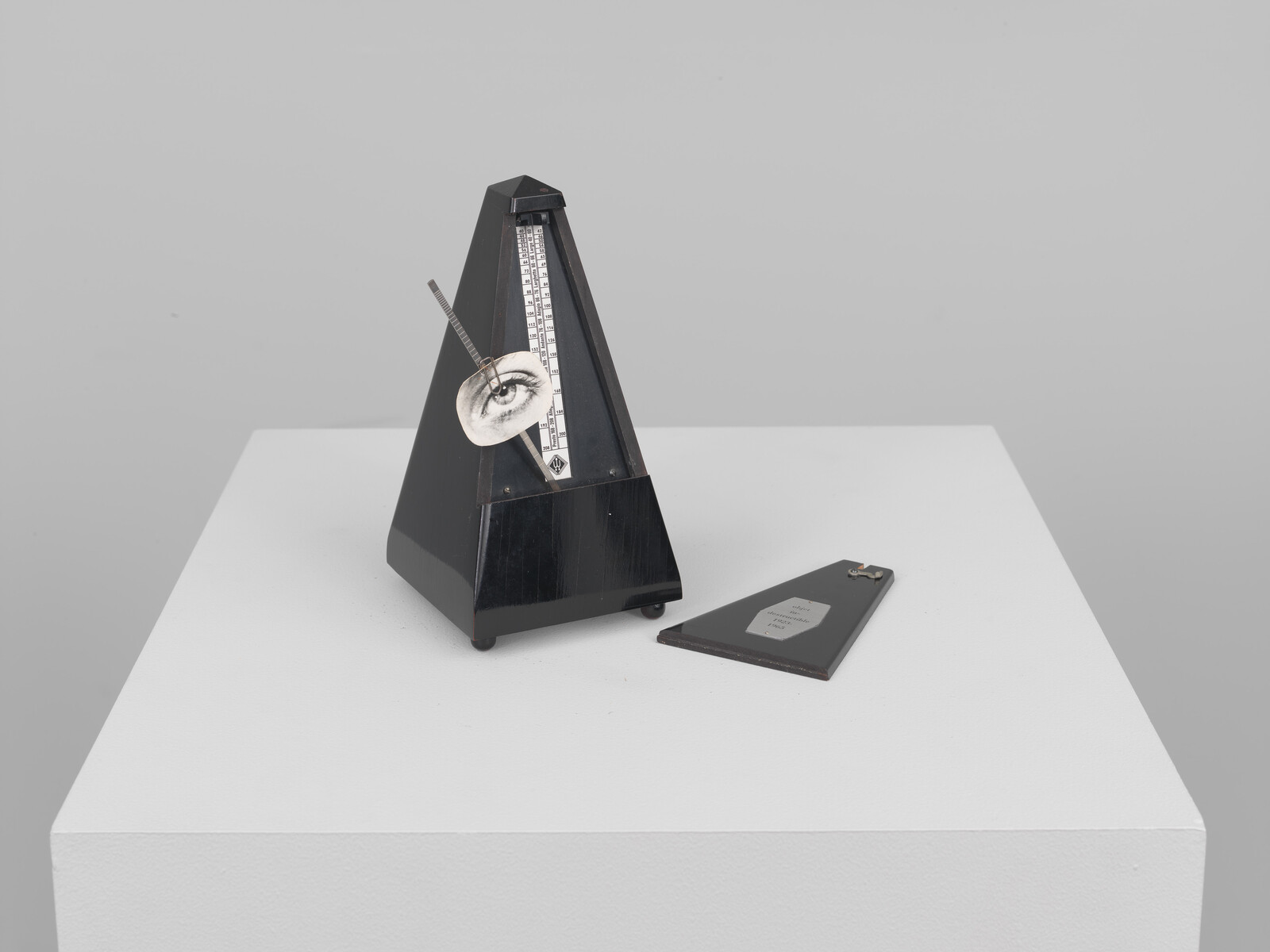 A black pyramid metronome with its pendulum showing a drawn eye resting on a white pedestal.