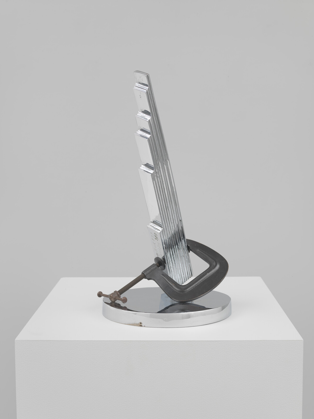Chrome stepped metal sculpture held at an angle by a large C-clamp on a round base.