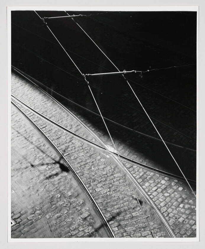 Black and white photograph depicting a close-up view of tram tracks intersecting over cobblestone pavement, with overhead wires casting shadows on the ground.