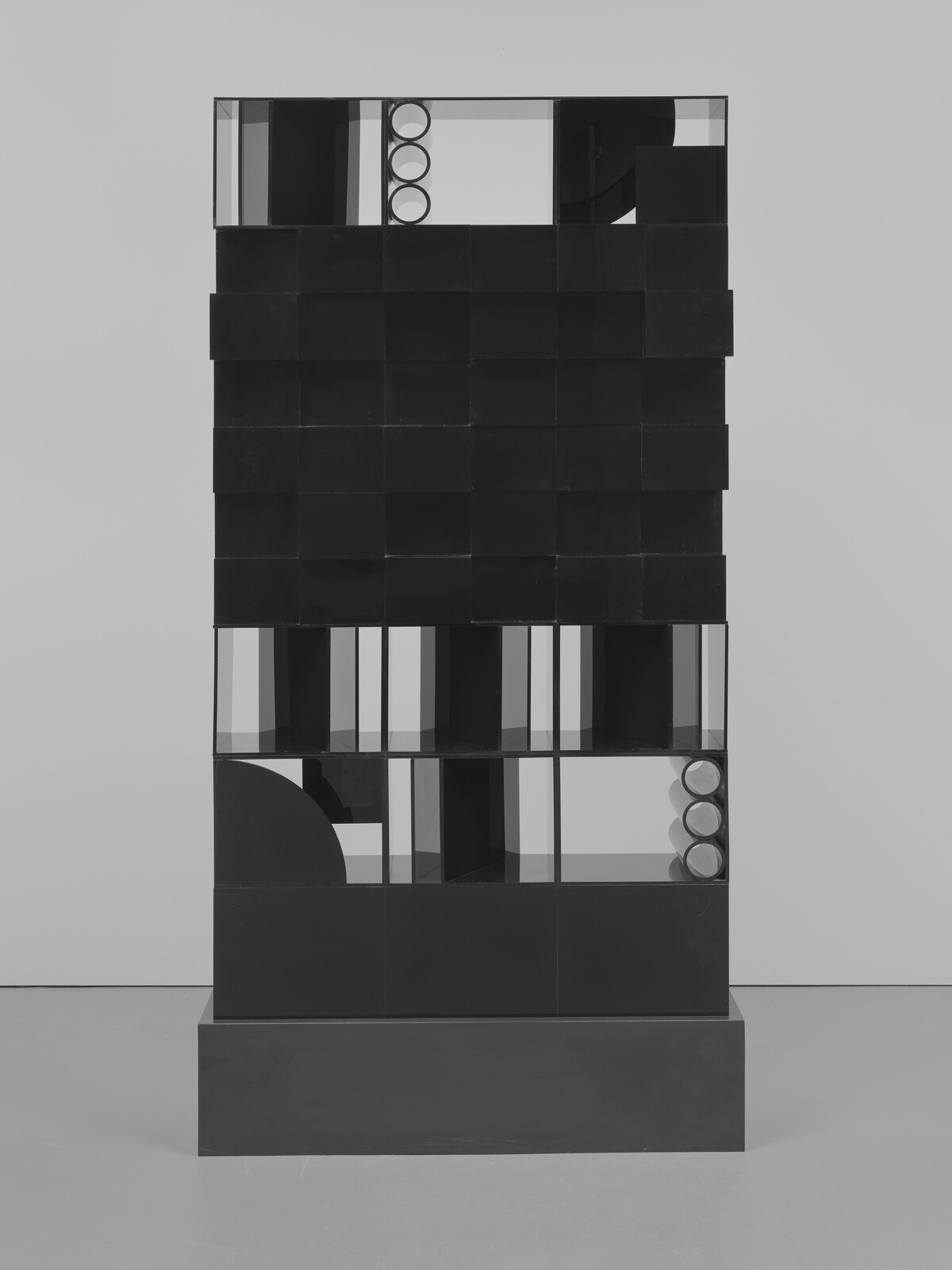 Tall black geometric sculpture with stacked square panels and open compartments showing three round tubes.