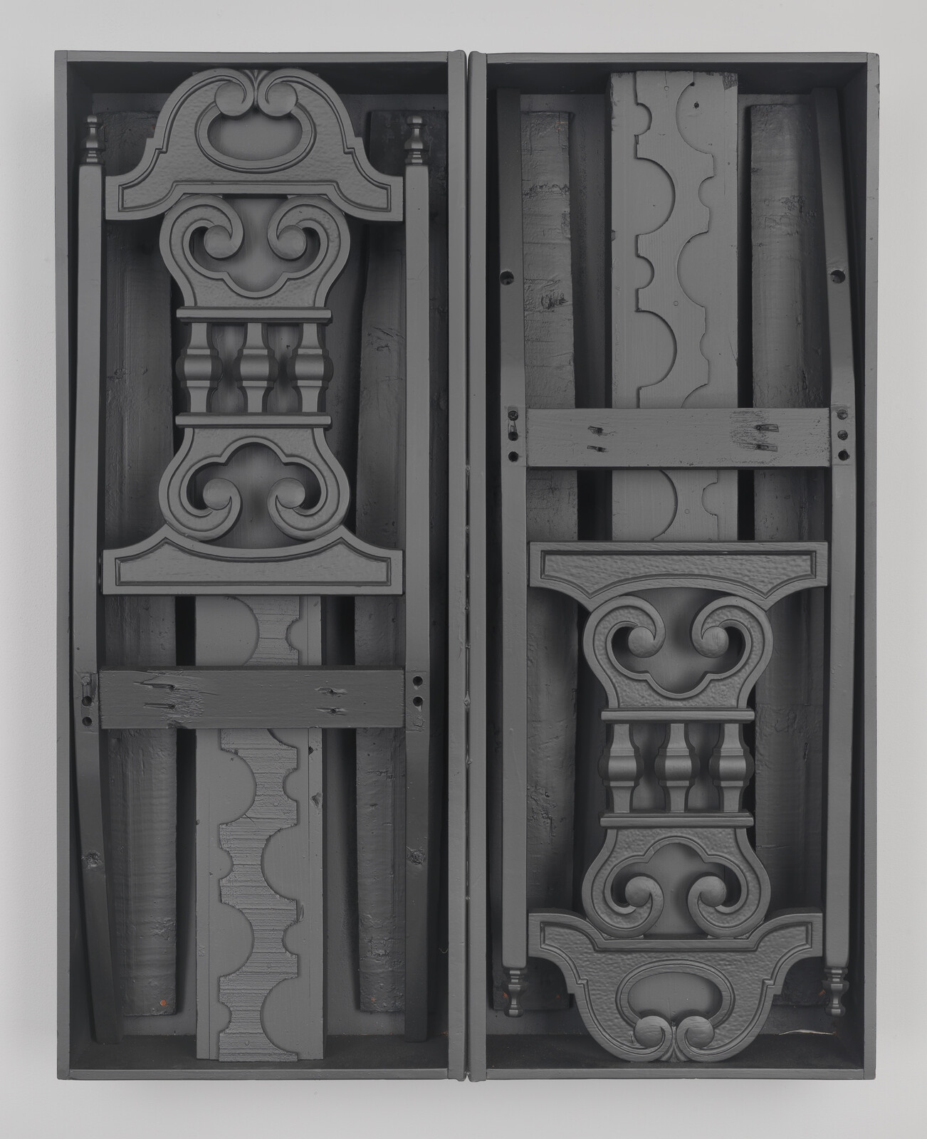 Two vertical wooden frames containing carved ornamental furniture parts with scrollwork and balusters.