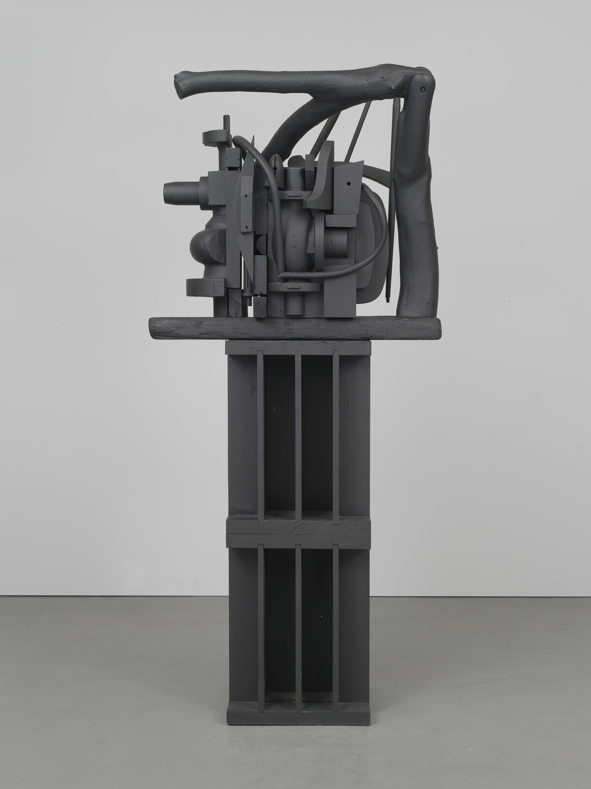 Dark gray abstract sculpture with mechanical elements on a tall pedestal against a plain background.