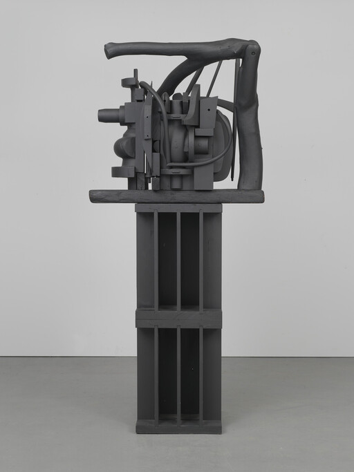 A dark gray abstract machine-like sculpture with pipes and wooden frame standing on a tall pedestal.